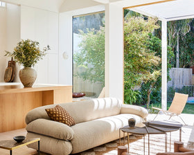 Modern living room with a beige sofa, wooden armchair, and large windows overlooking a garden.